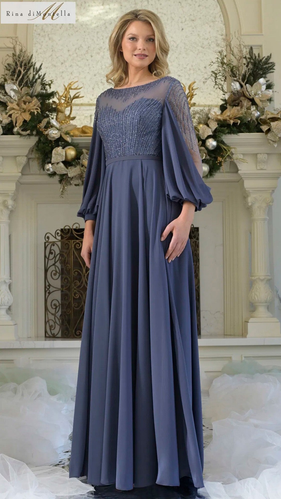 A woman stands indoors in front of a decorated white fireplace wearing the Rina Di Montella RD3374 long formal dress in slate blue, featuring chiffon fabric, sheer sleeves, a beaded bodice, and an elegant A-line skirt.